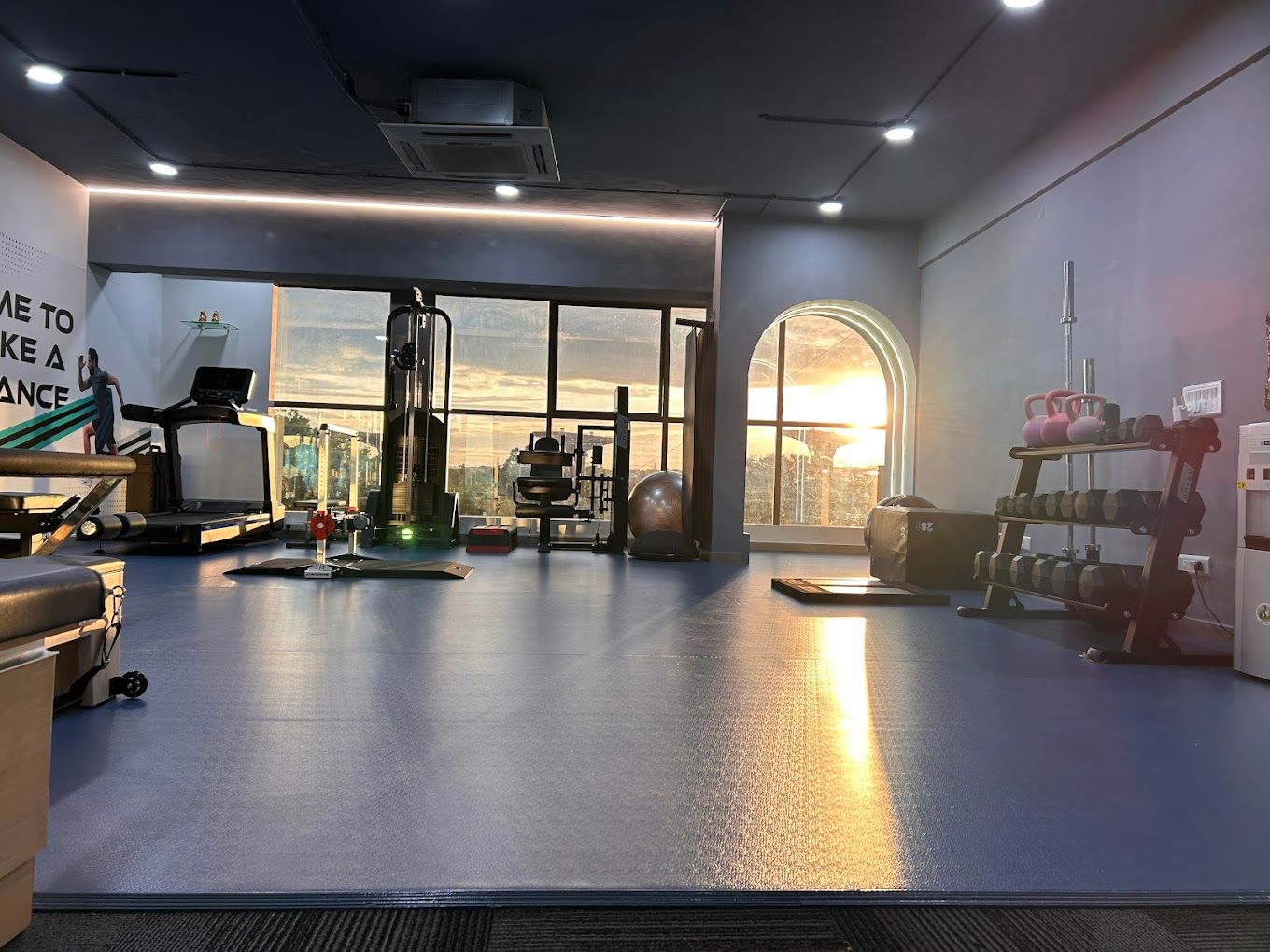 Gym floor with arched window, sunset light, treadmill and dumbbell rack